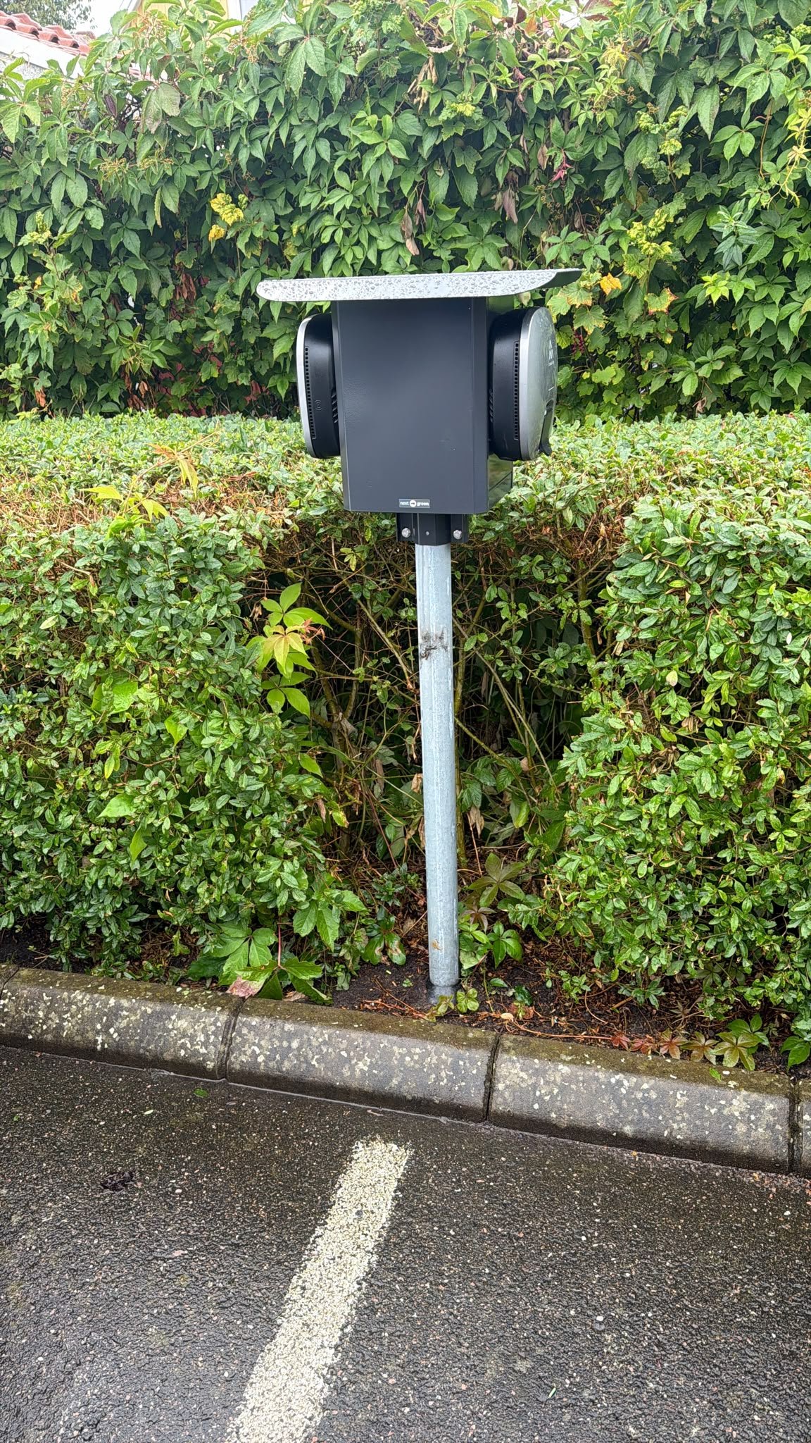 Stolpmonterad laddstation med två laddboxar och väderskydd vid parkeringsplats intill häck